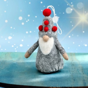 Könnte beinhalten: Ein grauer Filz-Zwergen-Anhänger mit einem roten Filzhut und roten Filzkugeln oben. Der Zwerg hat einen weißen Filzbart und hält eine Schnur.