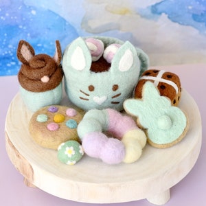 Puede incluir: Una taza de fieltro con forma de conejo llena de chocolate y malvaviscos, una galleta de fieltro con forma de conejo, una dona de fieltro, una galleta de fieltro con chispas, un pan de cruz de fieltro y un huevo de Pascua de fieltro sobre un soporte de madera.