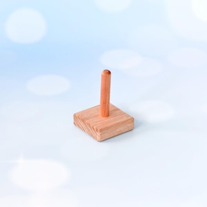 May include: A small, square wooden stand with a vertical, rounded dowel. The stand is made of light-colored wood and has a natural finish. The dowel is centered on the square base. The background is a soft blue with blurred white circles.