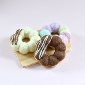 May include: Three felt doughnuts on a wooden chopping board. The doughnuts are in the colours of chocolate, white, and pastel purple and green.
