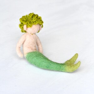 May include: A felt mermaid doll with green hair and a green tail. The doll is sitting on a white surface.