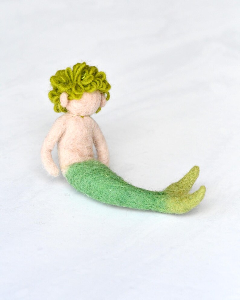 May include: A felt mermaid doll with green hair and a green tail. The doll is sitting on a white surface.