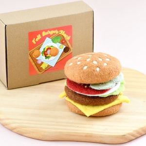 May include: A felt burger stack toy with a brown patty, yellow cheese, red tomato, green lettuce, and a tan bun with white sesame seeds. The burger is sitting on a wooden cutting board. The toy is packaged in a brown cardboard box with a label that says "Felt Burger Stack".