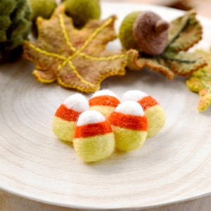May include: A group of handmade felt candy corn in yellow, orange, and white. The candy corn is displayed on a light wooden surface, with felt acorns and autumn leaves in the background. These felt decorations are ideal for autumn or Halloween.