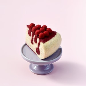 Può includere: Una fetta di cheesecake in feltro con guarnizione di frutti di bosco rossi, presentata su un piccolo alzatina lilla. La cheesecake ha una base bianca cremosa con salsa di frutti di bosco rosso scuro e frutti di bosco rossi. Lo sfondo è rosa tenue.