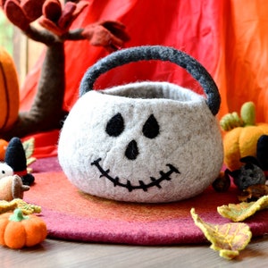 Op de afbeelding: Een witte viltpompoen-vormige trick-or-treat-emmer met een zwart viltgezicht en een gestikte glimlach. De emmer heeft een zwart vilt handvat en staat op een rode en oranje viltmat.