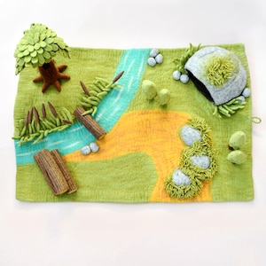 May include: A felt landscape scene with a river, tree, and rocks. The scene is primarily green and yellow, with a blue river and brown tree trunk. The rocks and tree have green accents. The scene includes two wooden logs.