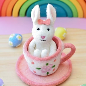 May include: A white felt bunny with a pink bow sits inside a pink teacup with a floral design. The teacup is on a pink saucer.