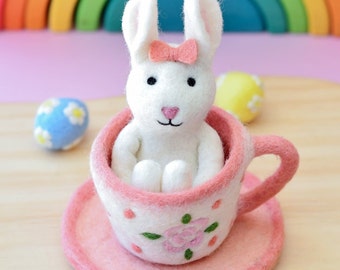 May include: A white felt bunny with a pink bow sits inside a pink teacup with a floral design. The teacup is on a pink saucer.