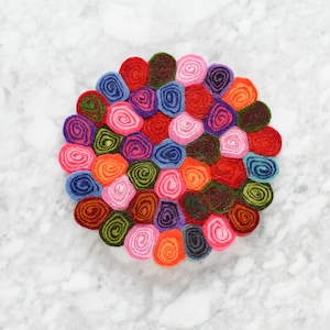 May include: A colourful felt coaster with a circular design made of many small, rolled felt circles in various shades of pink, red, purple, blue, green, and orange.