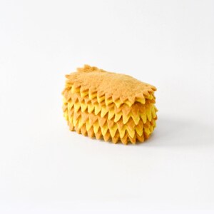 Puede incluir: Un objeto rectangular de textura suave con bordes dentados en capas en tonos alternos de amarillo dorado y marrón claro. La estructura está compuesta por múltiples segmentos de tela puntiagudos apilados que crean un aspecto denso.