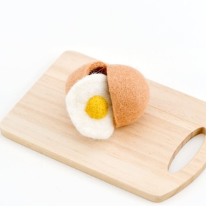 May include: A felt egg with a yolk and white inside, broken open and displayed on a wooden chopping board.