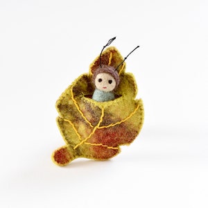 May include: A handmade felt leaf with a small figure inside. The leaf is primarily yellow and green with brown and red accents, and yellow stitching. The figure has a brown hat, a blue shirt, and black antennae.