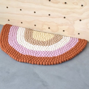May include: A semi-circular rug with a rainbow design. The rug features a gradient of colors, including rust, pink, white, tan, and yellow. The rug is made of small, round, textured balls. The rug is placed on a gray surface.