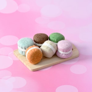 Peut inclure: Six macarons en feutre de différentes couleurs, dont le marron, le vert, le bleu clair, le beige et le rose, disposés sur une planche à découper en bois. Les macarons sont sur un fond rose avec des cercles blancs.