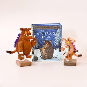 Puede incluir: Un libro infantil titulado "The Gruffalo's Child" está flanqueado por dos figuras de fieltro del Gruffalo sobre bases de madera. Las figuras son marrones con detalles morados. La portada del libro muestra una escena invernal.