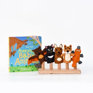 Può includere: Un libro per bambini intitolato "Australian Baby Animals" accanto a un set di cinque burattini a dito in feltro raffiguranti animali australiani. I burattini sono marroni, neri e arancioni, montati su tasselli di legno e una base di legno.