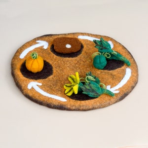 Könnte beinhalten: Eine Filzspielmatte mit drei braunen Filzpatches, jeder mit einem anderen Filzgegenstand. Die Gegenstände sind ein kleiner orangefarbener Kürbis, eine grüne Filzkürbis und eine grüne und gelbe Filzblume. Weiße Pfeile sind um den Rand der Matte gezeichnet.