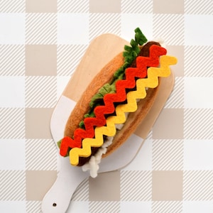 Puede incluir: Un perrito caliente de fieltro en una tabla de cortar de madera con coberturas de fieltro amarillo, rojo y verde. La tabla de cortar está sobre un fondo de cuadros blancos y marrones.