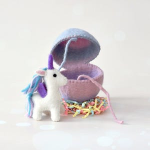May include: A white felt unicorn with blue and purple mane stands beside a felt egg. The egg is light blue and pink, with a pink string. The unicorn is standing on a bed of colourful paper strips.