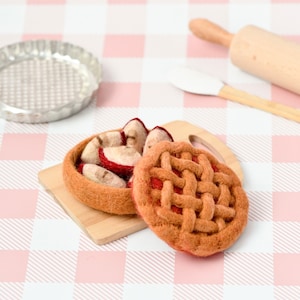 May include: Two felt apple pies on a wooden cutting board. The pie crusts are brown and the filling is red. The pies are on a pink and white checkered tablecloth.