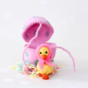 Puede incluir: Decoración de Pascua hecha a mano. Un patito amarillo con un gorro rosa junto a una cáscara de huevo rosa adornada con pequeñas flores azules y verdes. El patito está sobre confeti de papel colorido. Una encantadora decoración de Pascua.