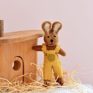 May include: A brown felt bunny wearing yellow overalls with a green patch pocket. The bunny is standing next to a wooden playhouse.