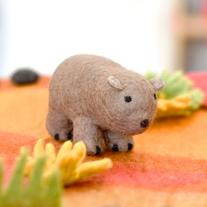 May include: A brown felt toy animal resembling a capybara. The toy has black eyes and a small pink nose. It is standing on a textured orange surface.