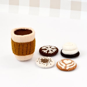 Puede incluir: Conjunto de juguetes de fieltro de una taza de café marrón con un borde blanco y una tapa marrón, y cuatro galletas de fieltro con diferentes diseños. Las galletas son una galleta de copo de nieve marrón, una galleta blanca con chispas marrones, una galleta blanca con glaseado marrón y un diseño de corazón, y una galleta blanca con glaseado marrón y un diseño de remolino.