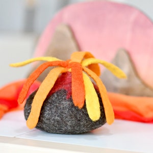 May include: A felt volcano toy with a gray base and orange and yellow felt flames. The volcano is sitting on a white surface.