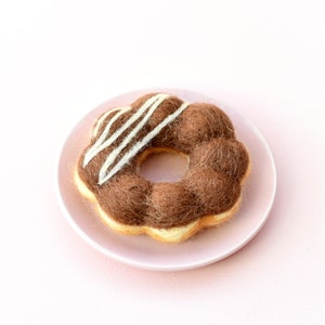 May include: A brown and white felted doughnut with white icing on a pink plate.