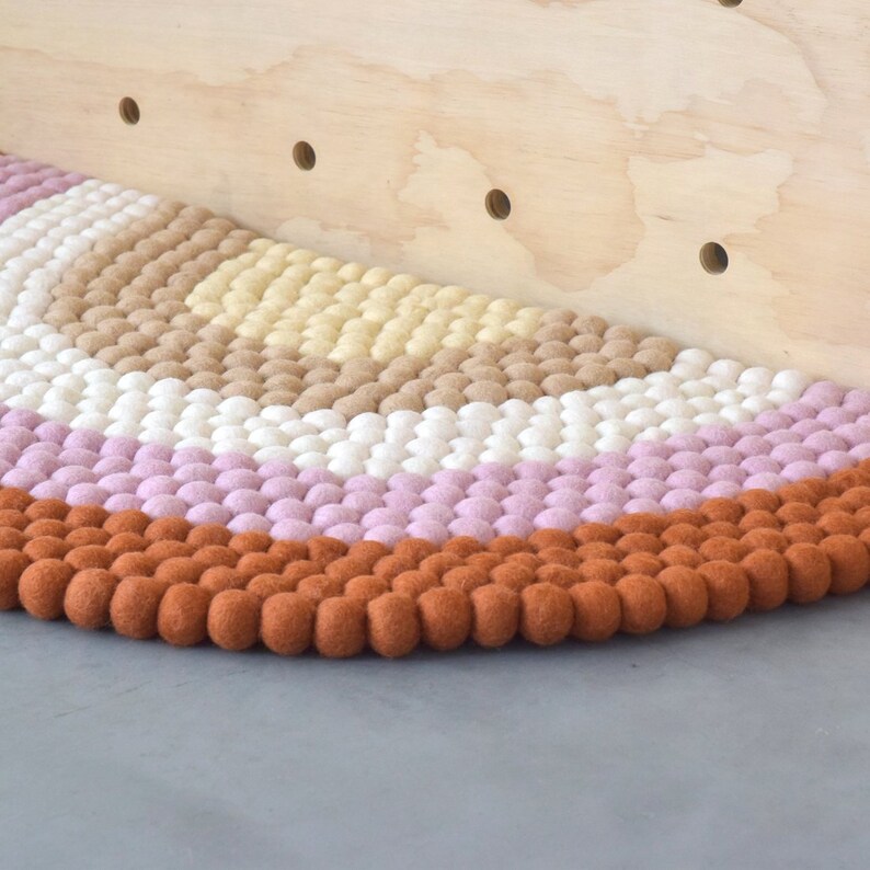 May include: A colorful, semi-circular rug with a textured, felted design. The rug features bands of various colors, including brown, white, pink, and orange. The rug is placed against a light-colored wooden surface.