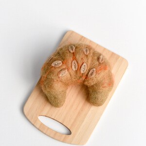 May include: A brown felt croissant with orange stitching and beige felt leaves on a wooden cutting board.