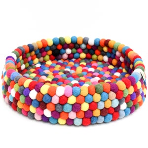 May include: A round pet bed crafted from colorful felt balls. The bed features a vibrant mix of red, orange, yellow, green, blue, purple, and white balls, creating a textured surface. The bed's design is suitable for small pets.