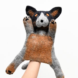 May include: A gray and brown felt hand puppet of a dog with black ears and orange eyes. The puppet is being held up by a hand.