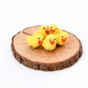 May include: Five yellow felt chicks with orange beaks and black eyes are arranged on a round wooden slice. The chicks are clustered together, creating a cheerful, springtime theme. The wooden slice has a natural, rustic appearance.