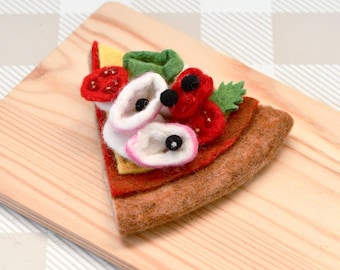 May include: A felt pizza slice with red tomato, white cheese, green basil, and black olives on a wooden cutting board.