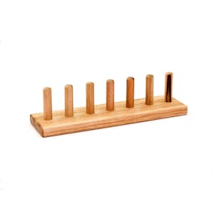 May include: A wooden plate rack with seven vertical pegs. The rack is made of light-colored wood and has a rectangular base. The pegs are evenly spaced and cylindrical. One peg is darker than the others. The background is white.