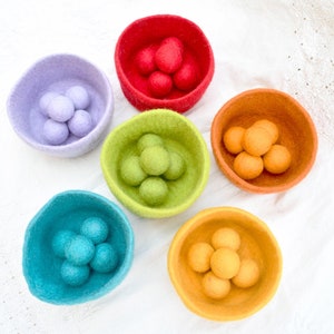 May include: Six felt bowls in different colours, each filled with felt balls of the same colour. The bowls are arranged in a circle on a white surface. The colours of the bowls are red, purple, green, orange, blue, and yellow. The colours of the felt balls are red, purple, green, orange, blue, and yellow.
