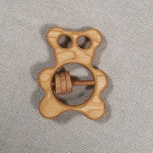 May include: Wooden bear rattle toy with a spinning wooden cylinder in the center. The toy is made of natural wood and has a light brown color.