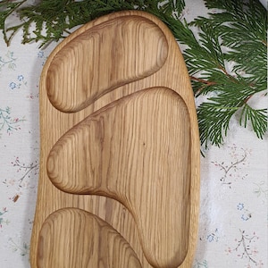 May include: A wooden serving tray with three separate compartments, each shaped like an oval. The tray is made of light brown wood with a natural finish.