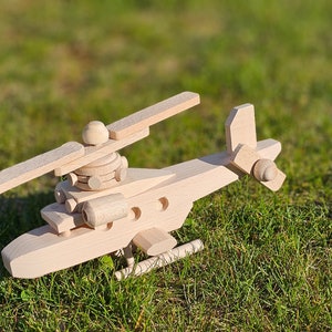 Könnte beinhalten: Ein hölzerner Spielzeug-Hubschrauber mit hellbrauner Oberfläche. Der Hubschrauber hat einen Rotor, einen Heckrotor und einen Rumpf mit einem kleinen Fenster. Das Spielzeug sitzt auf einem Bett aus grünem Gras.