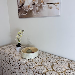 Op de afbeelding: Decoratieve scène met een wit kanten tafelkleed met een bloemmotief. Een kleine orchidee in pot en een decoratieve kom met een steen en rode bessen staan op de tafel. Een schilderij van witte orchideeën hangt aan de muur.