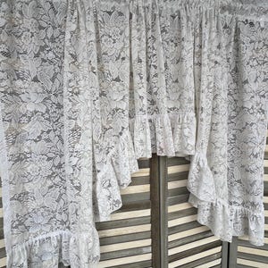 May include: White lace curtain with a floral pattern and ruffled edges. The curtain is hanging in front of a wooden folding screen. The lace is delicate and the design is intricate, creating a soft, romantic look.