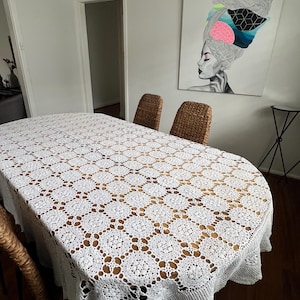 May include: A white, oval crochet tablecloth with a detailed lace pattern covers a wooden dining table. The design features repeating floral motifs. Two woven wicker chairs are visible. A painting of a woman with a colourful headpiece hangs on the wall.