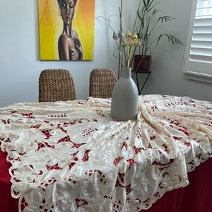 May include: A dining table set with a red tablecloth and a cream-coloured, intricately patterned overlay. A grey vase holds decorative flowers. A painting of a woman with a head wrap hangs on the wall.