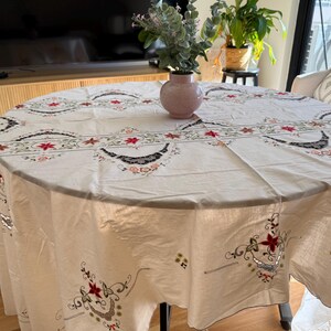 May include: A round off-white tablecloth featuring intricate embroidery with red floral motifs, green leafy vines, and black crescent-shaped patterns. The fabric drapes over a circular table, which is topped with a small pink vase containing greenery.