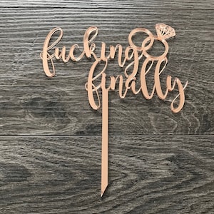 Könnte beinhalten: Tortenaufsatz aus roségoldfarbenem Acryl mit dem Text "fucking finally" und einem Paar stilisierter Ringe mit einem Diamanten.