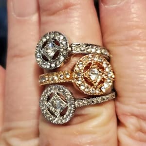 May include: Three silver, gold, and rose gold rings with clear stones set in a square shape. The rings are stacked on a finger.
