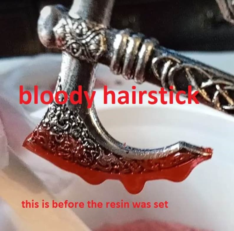 May include: A silver axe-shaped hair stick with intricate details, dripping with red resin. The text "bloody hairstick" is written in red letters above the hair stick. The text "this is before the resin was set" is written below the hair stick.